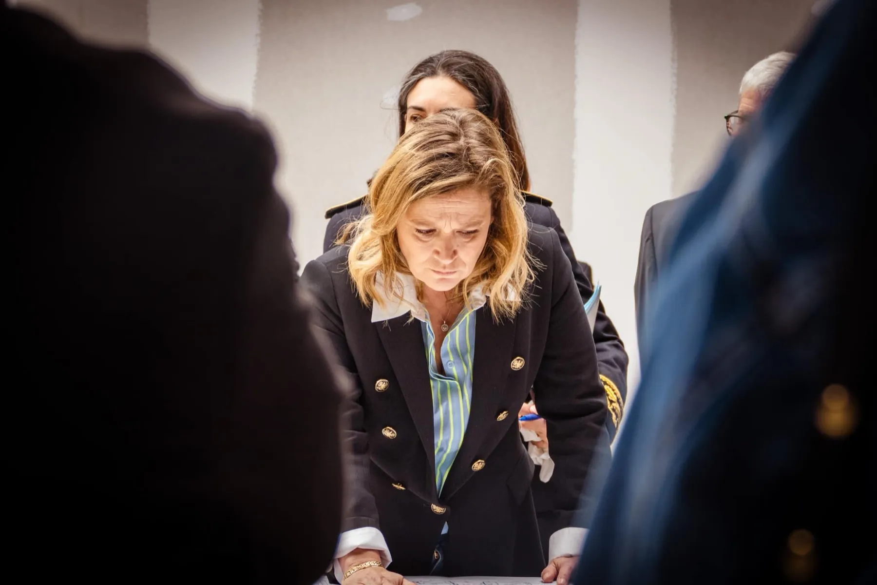 Femme blonde en blazer penchée sur des documents, entourée de personnes