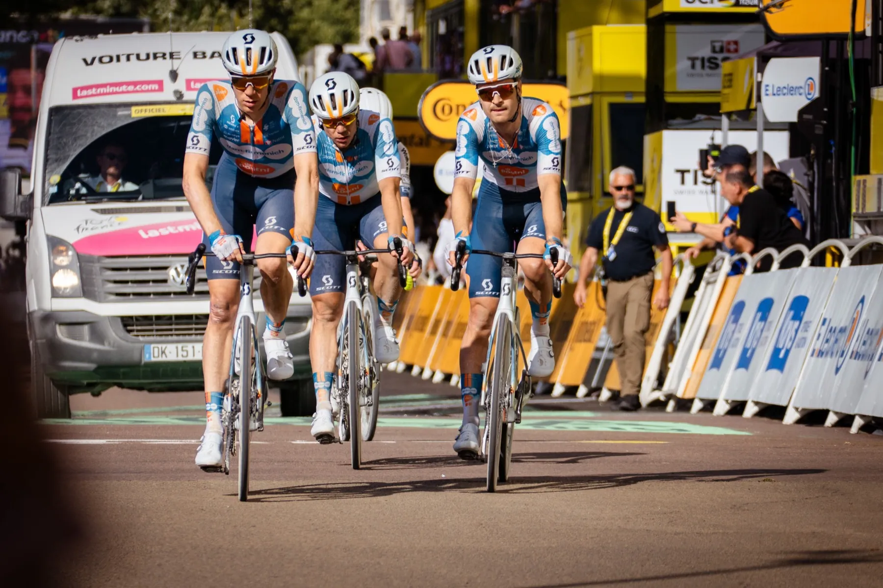 Trois coureurs cyclistes en course, véhicule d'équipe et barrières en arrière-plan